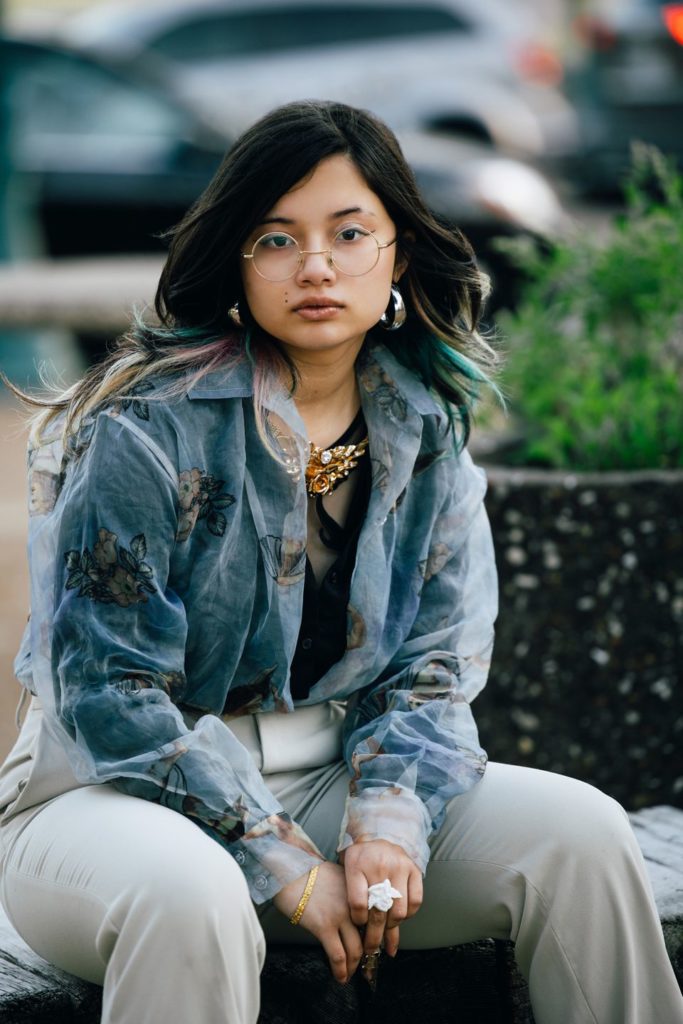 St. Louis Senior from Maplewood Richmond Heights High School is sitting on a bench in University City. Her dark hair is blowing in the wind to reveal blue flyaway highlights. she is wearing glasses, a vintage gold necklace, and sheer blue blouse.
