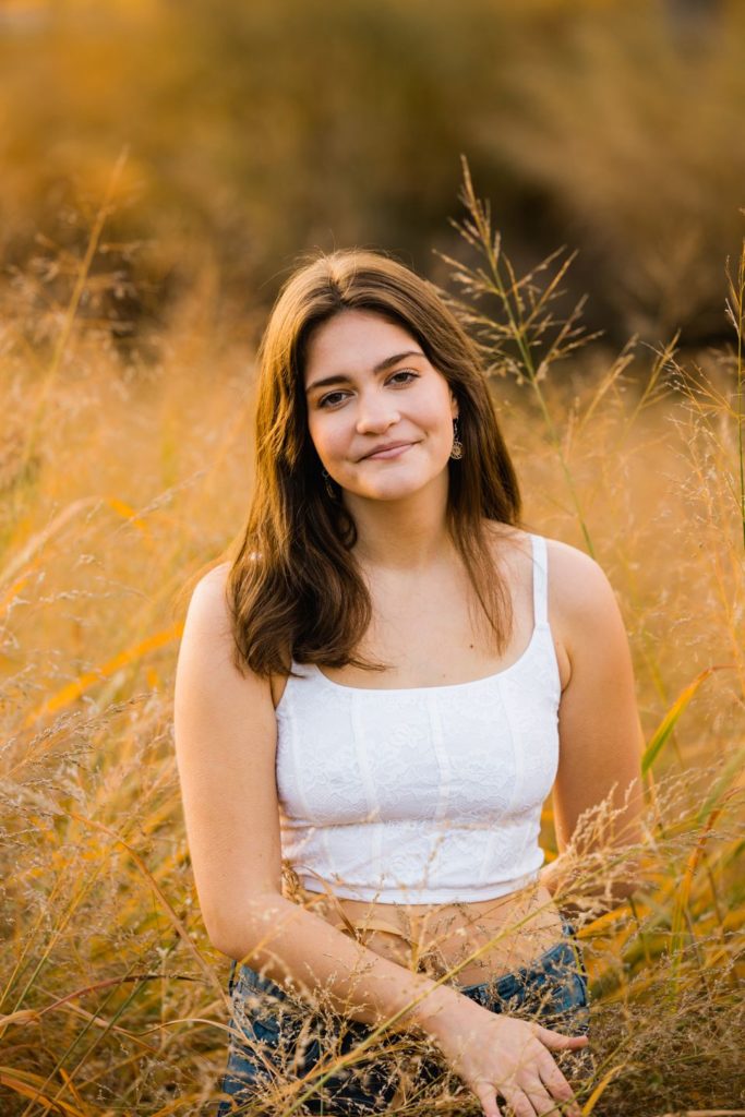 Dark haired senior from Webster Groves High School stand in tall yellow grass. She is wearing a white tank top and looking directly into the camera.